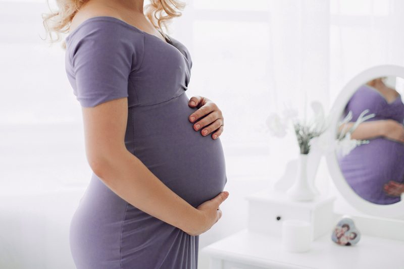 вагітна жінка в фіолетовому платті тримає живіт перед дзеркалом